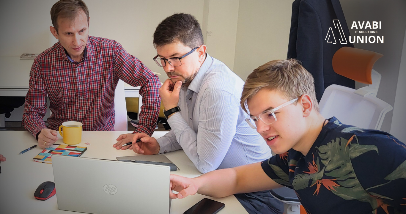 three men sitting at a table, looking at a computer monitor, discussing something.